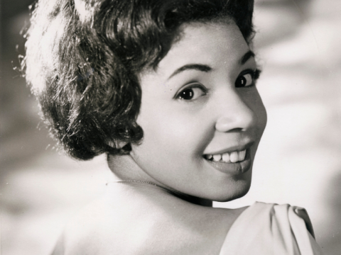 A close up, black and white photograph of musician Shirley Bassey smiling at the camera over her shoulder.